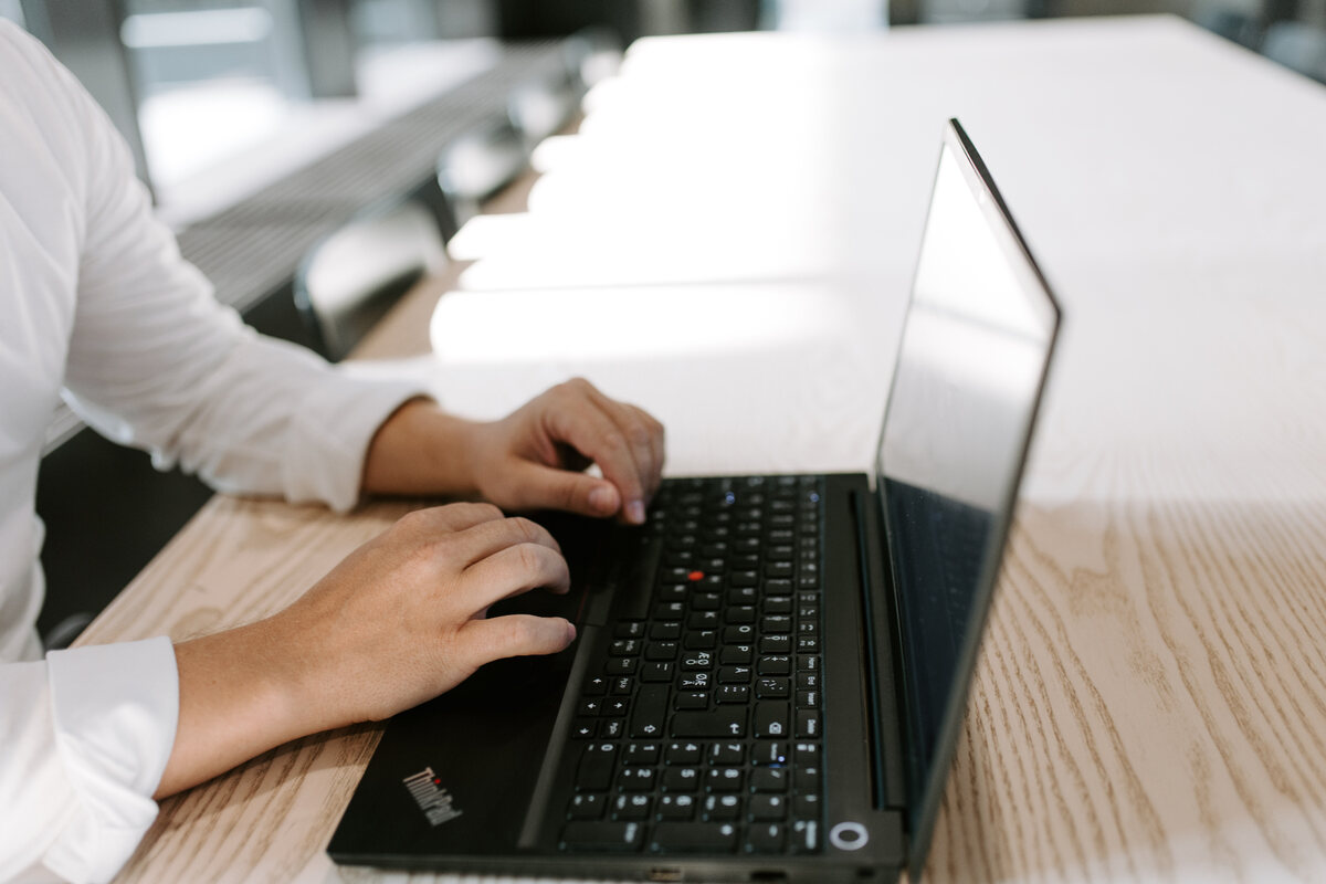 Lähikuva kahdesta kädestä, jotka kirjoittavat mustan ThinkPad-kannettavan näppäimistöllä vaalealla puupöydällä neuvotteluhuoneessa.