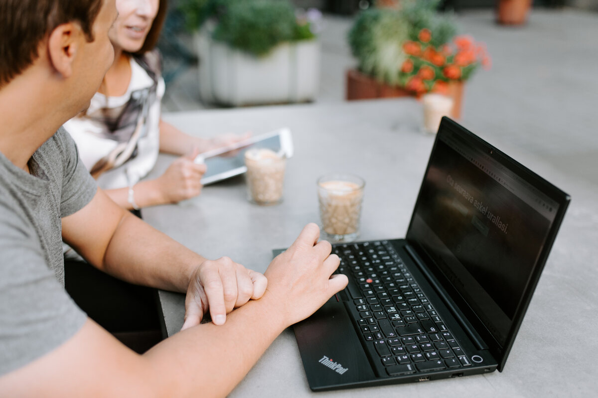 Mies ja nainen työskentelevät yhdessä ThinkPad-kannettavan ja tabletin ääressä ulkoterassilla, pöydällä kahvilaseja ja taustalla viherkasveja.