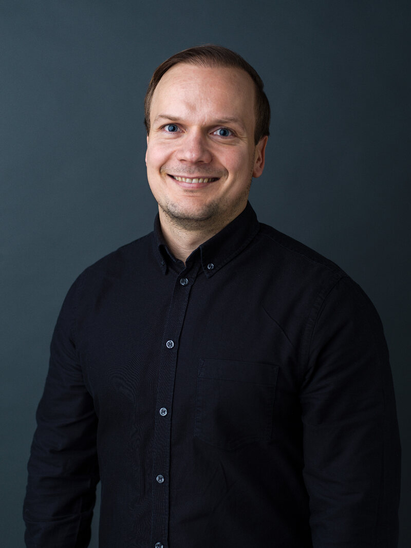 Hymyilevä tummaan pikeepaitaan pukeutunut mies studiomuotokuvassa tummansinistä taustaa vasten.
